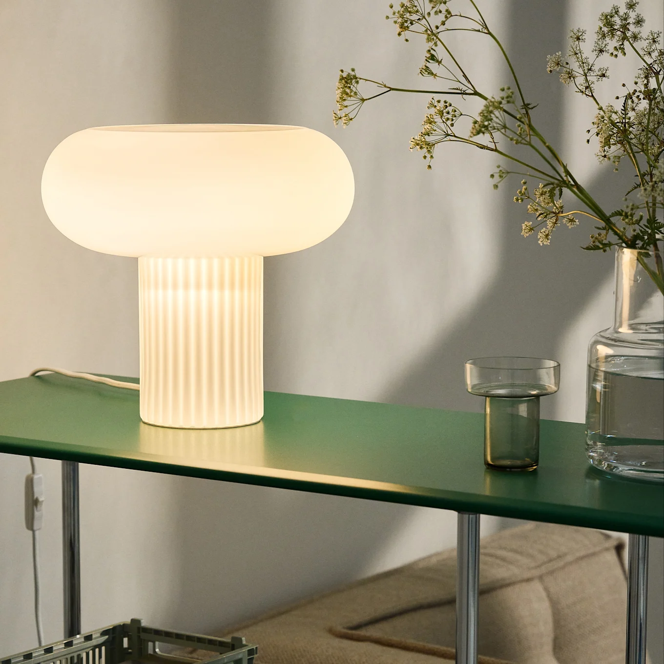 Lámpara de mesa luminosa con forma de seta y base acanalada en un estante verde oscuro, con flores en un jarrón.