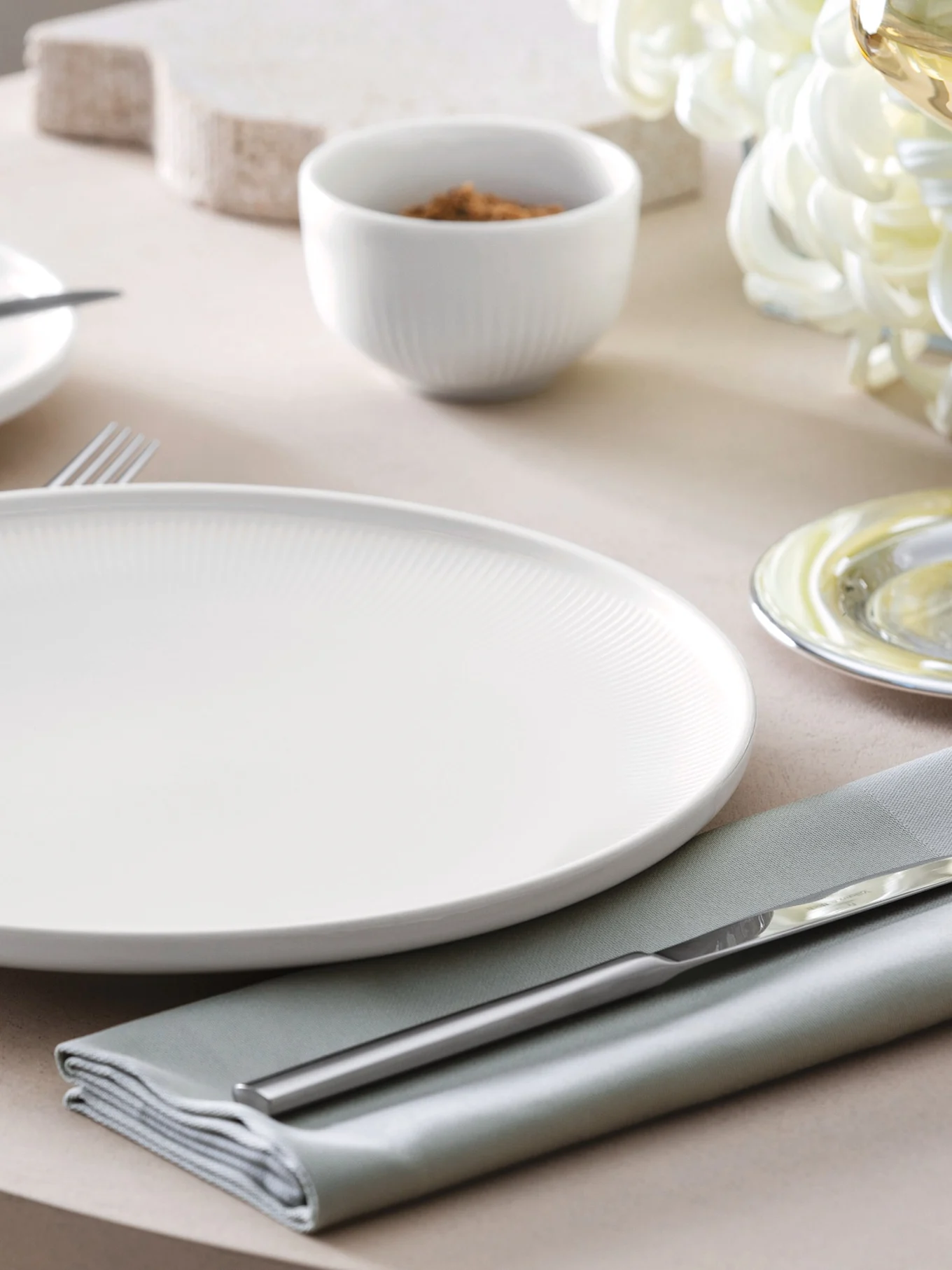 Un plato llano blanco estriado, servilleta verde salvia y cubiertos plateados sobre una mesa con un tazón pequeño al fondo.