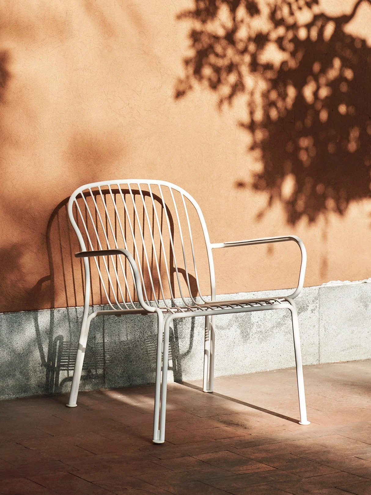 Silla de exterior de metal blanco con lamas contra una pared naranja con sombras de árboles en un día soleado.