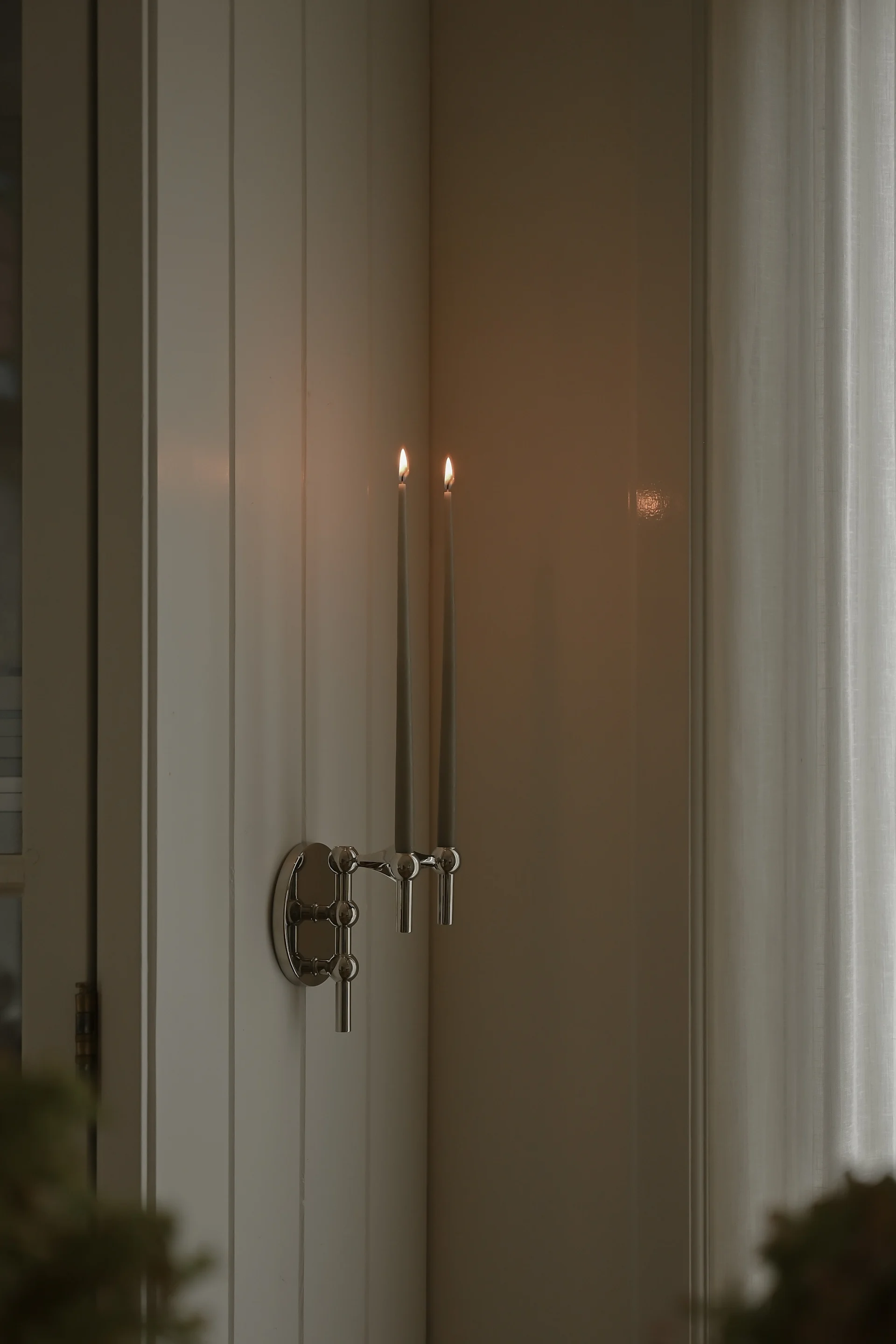 Un candelabro de pared plateado sostiene dos velas grises encendidas en una pared con paneles junto a una cortina transparente.