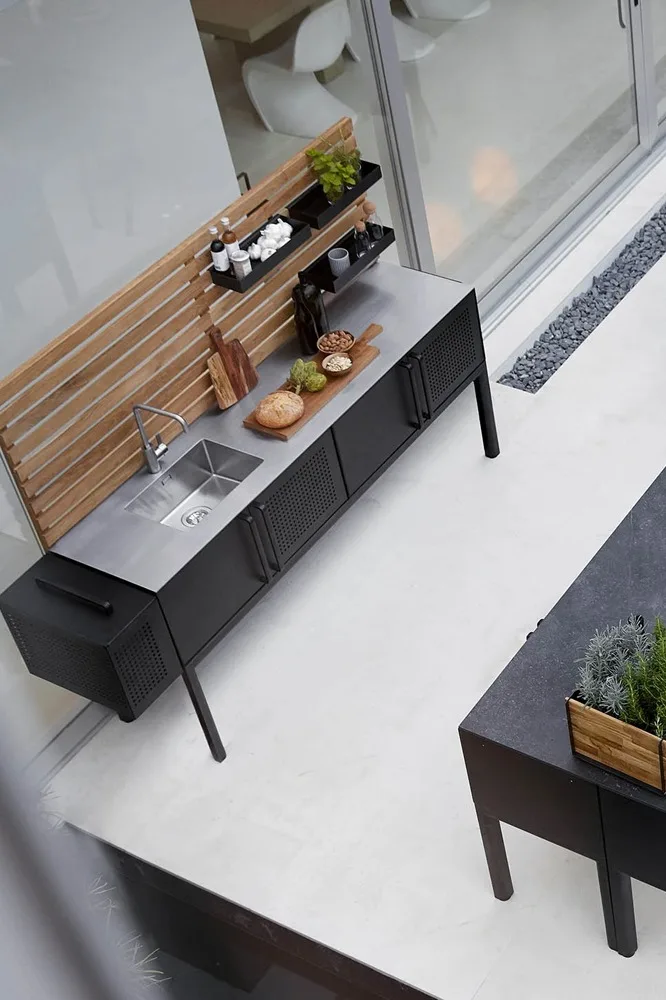 Moderna cocina exterior con unidad negra, fregadero de acero inoxidable, panel de madera, comida en tabla de cortar y jardinera de hierbas.