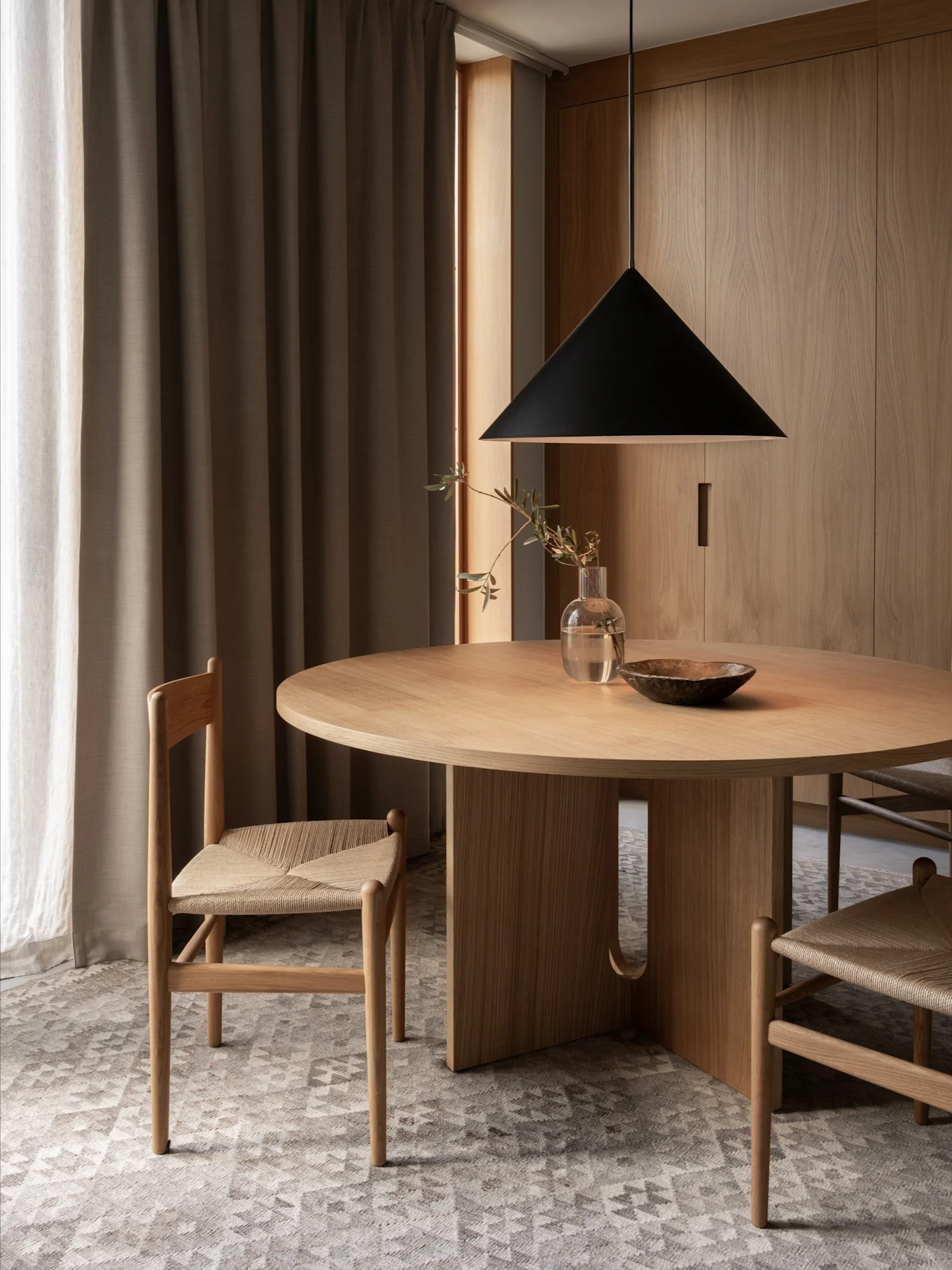 Un comedor minimalista con una mesa redonda de madera, dos sillas con asientos de mimbre, una lámpara colgante negra y paredes de madera.