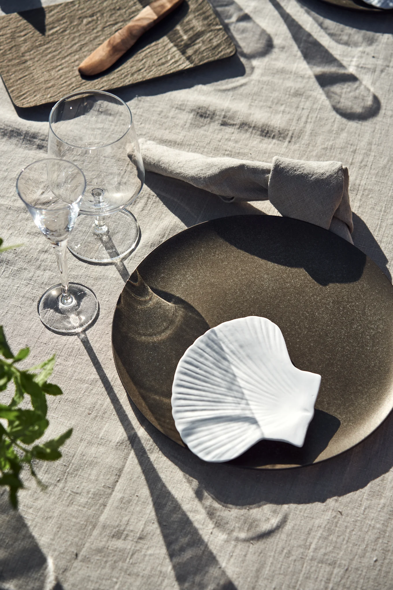 Un servicio de mesa individual con un plato llano oscuro, un plato blanco en forma de concha, dos copas de vino y una servilleta de lino sobre un mantel claro, con sombras pronunciadas.