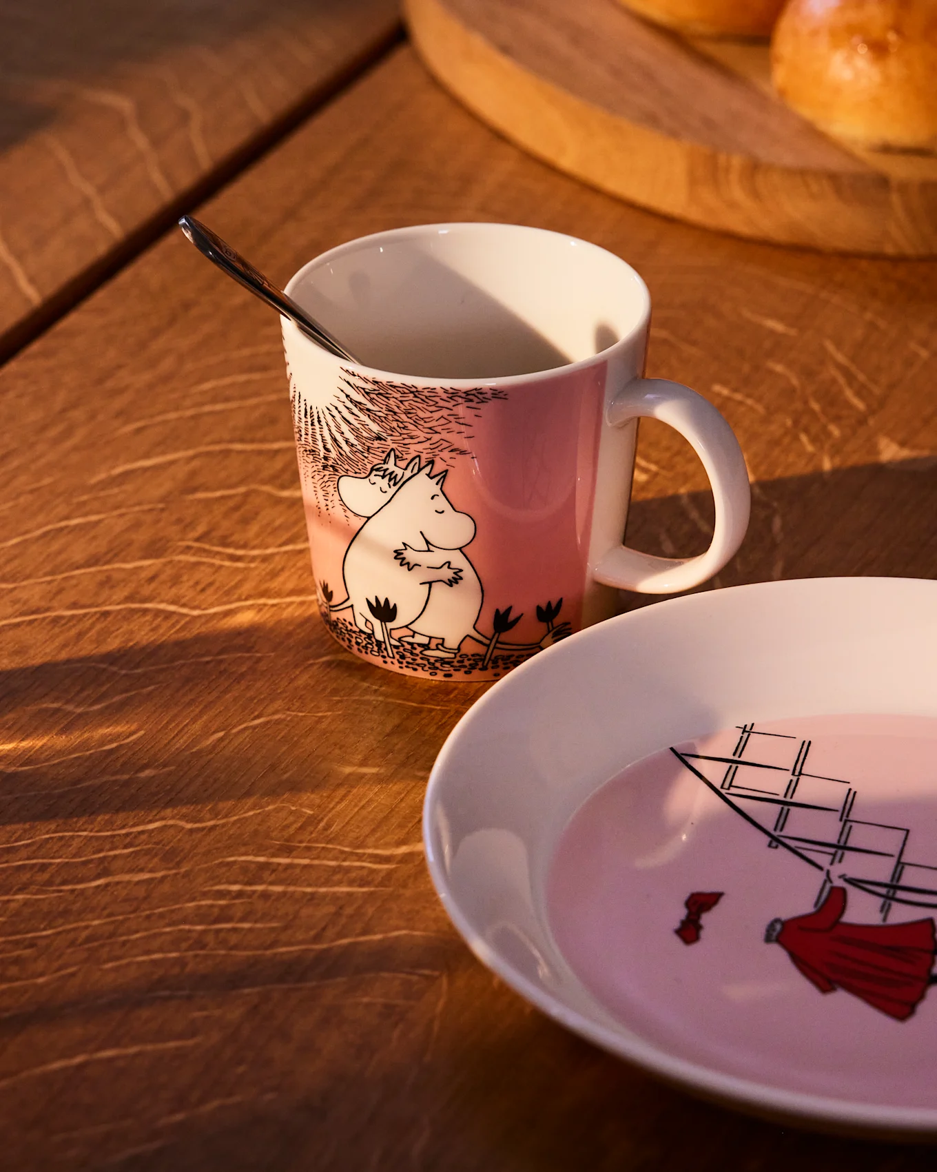 Taza Moomin rosa con Moomintroll y Snorkmaiden, junto a un plato a juego con un velero, sobre una mesa de madera.