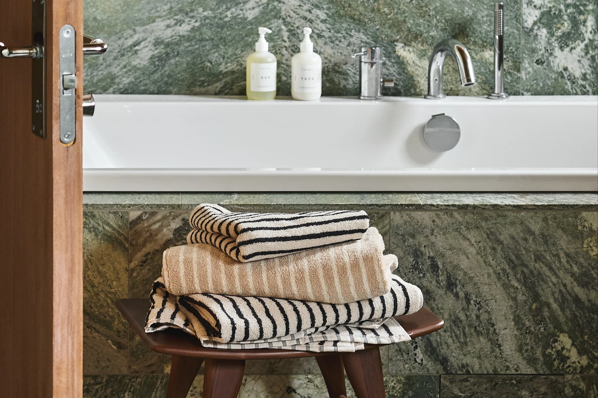 Una pila de toallas beige y rayadas sobre un taburete de madera en un baño con bañera blanca y paredes de mármol verde.