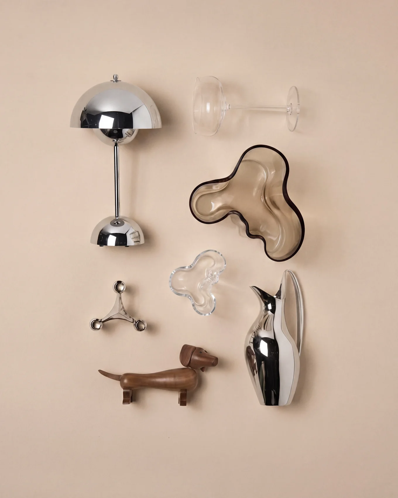 Decoración moderna: lámpara seta cromada, copa de vino, cuenco ondulado marrón, candelabro plateado, perro salchicha de madera, jarra plateada.