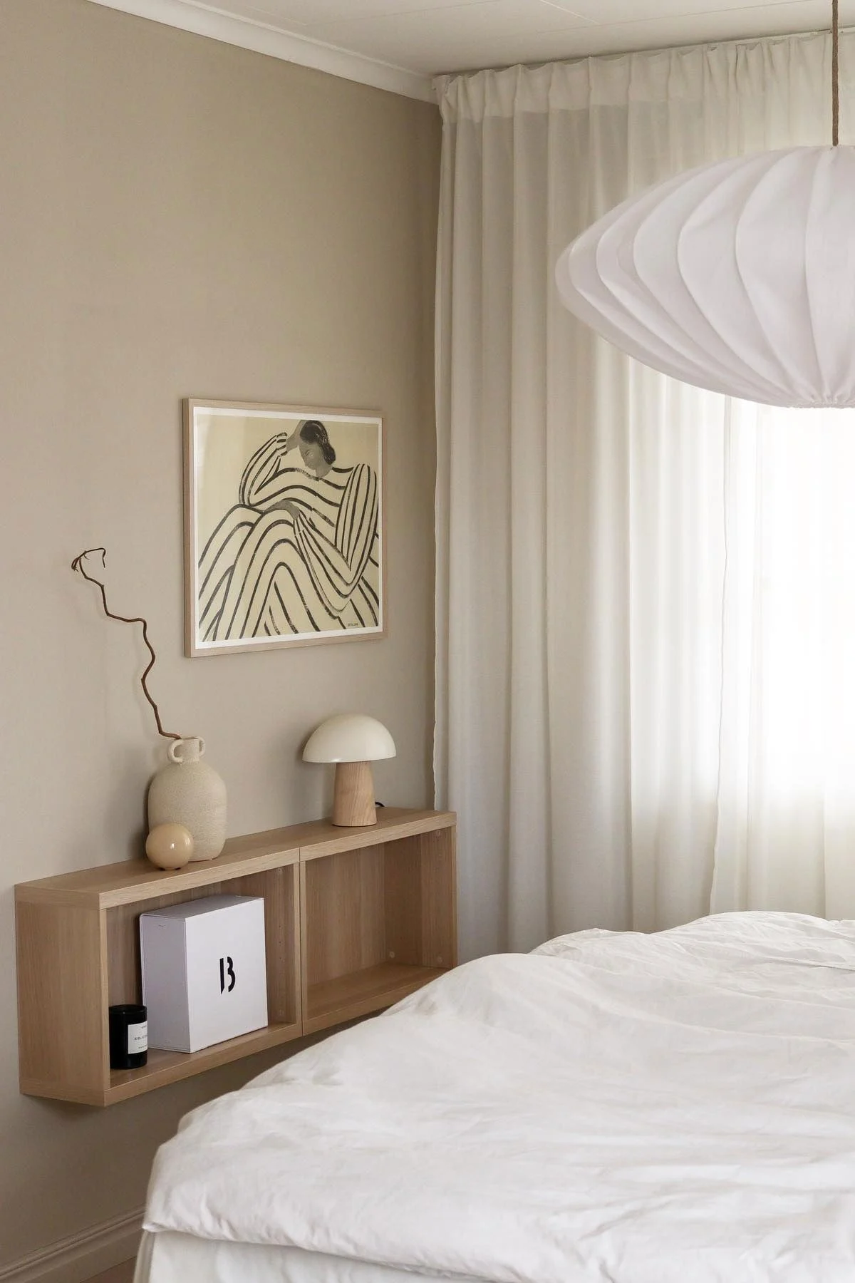 Dormitorio minimalista con ropa de cama blanca, estante flotante de madera con decoración, arte abstracto, cortinas transparentes y lámpara colgante plisada.