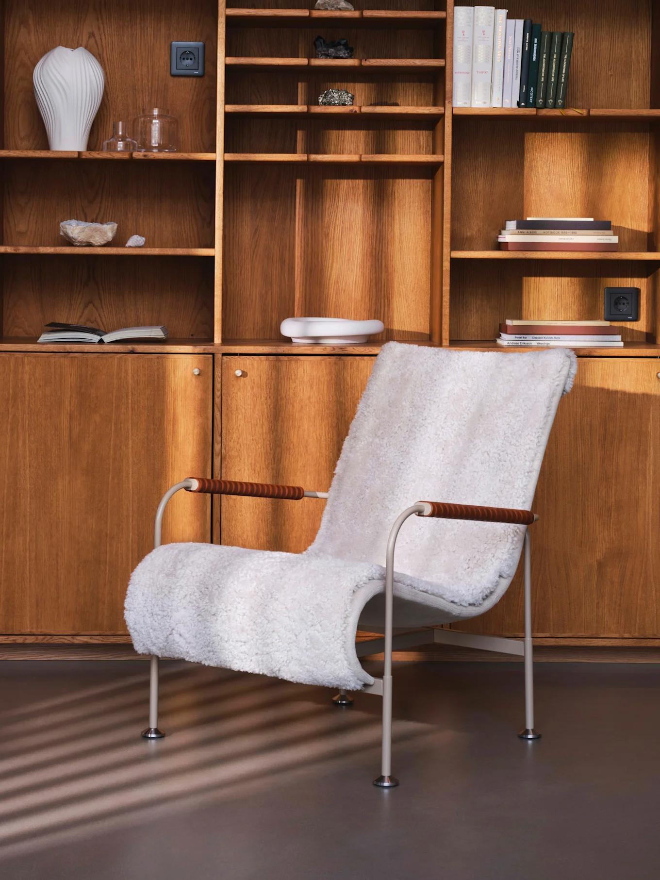 Sillón blanco de piel sintética frente a un mueble de pared de madera con estantes, libros, jarrones y minerales.