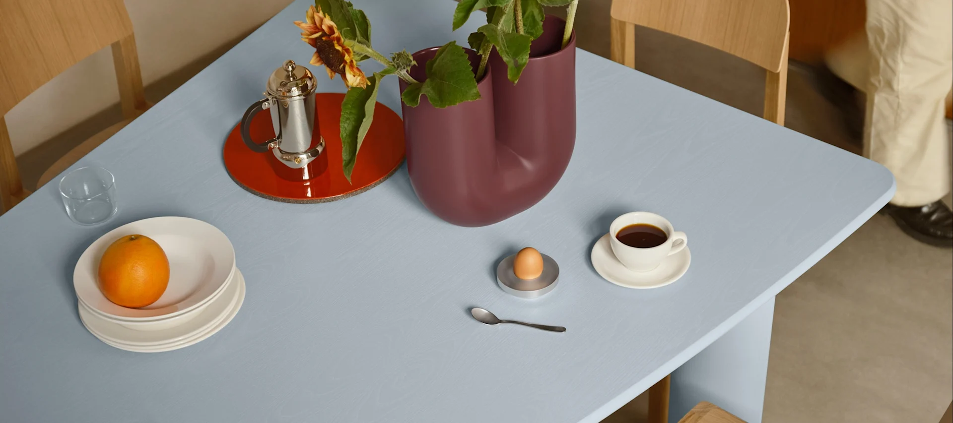 Mesa azul claro con desayuno: naranja en platos, café, huevo en huevera y un jarrón con girasoles.