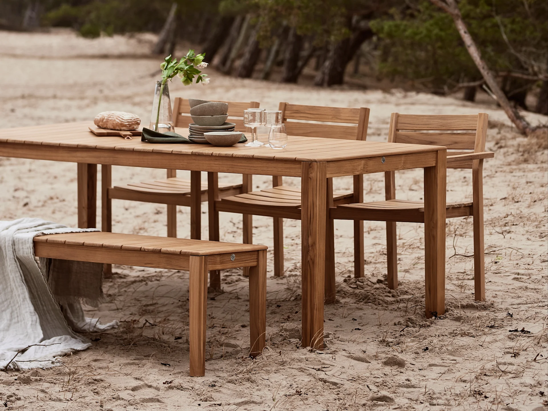 Juego de comedor exterior de madera con mesa, sillas y banco, decorado con vajilla y manta en una playa de arena.
