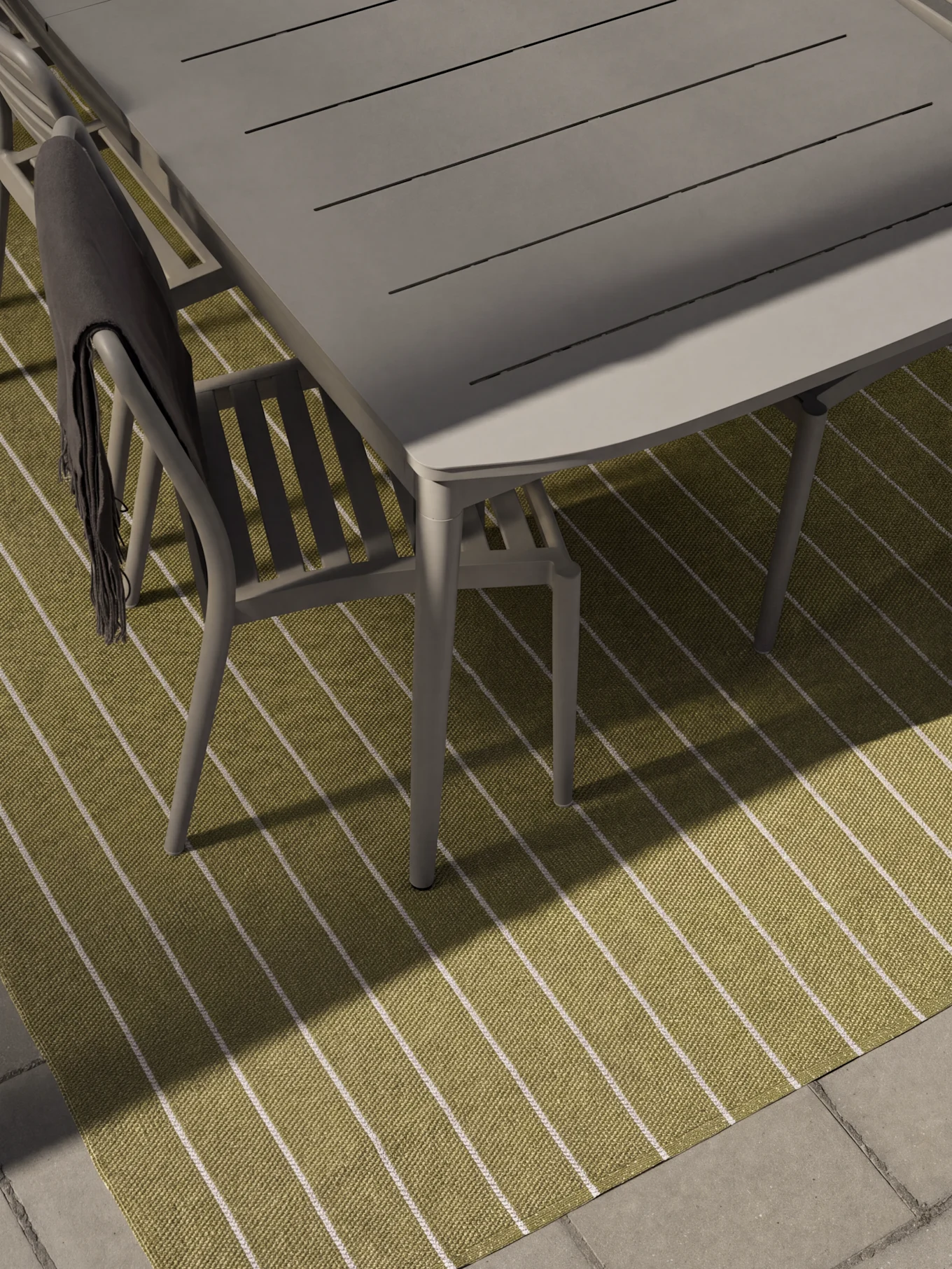 Mesa y sillas de comedor grises para exterior sobre una alfombra de rayas verdes y blancas. Una manta gris oscuro cuelga de una silla.