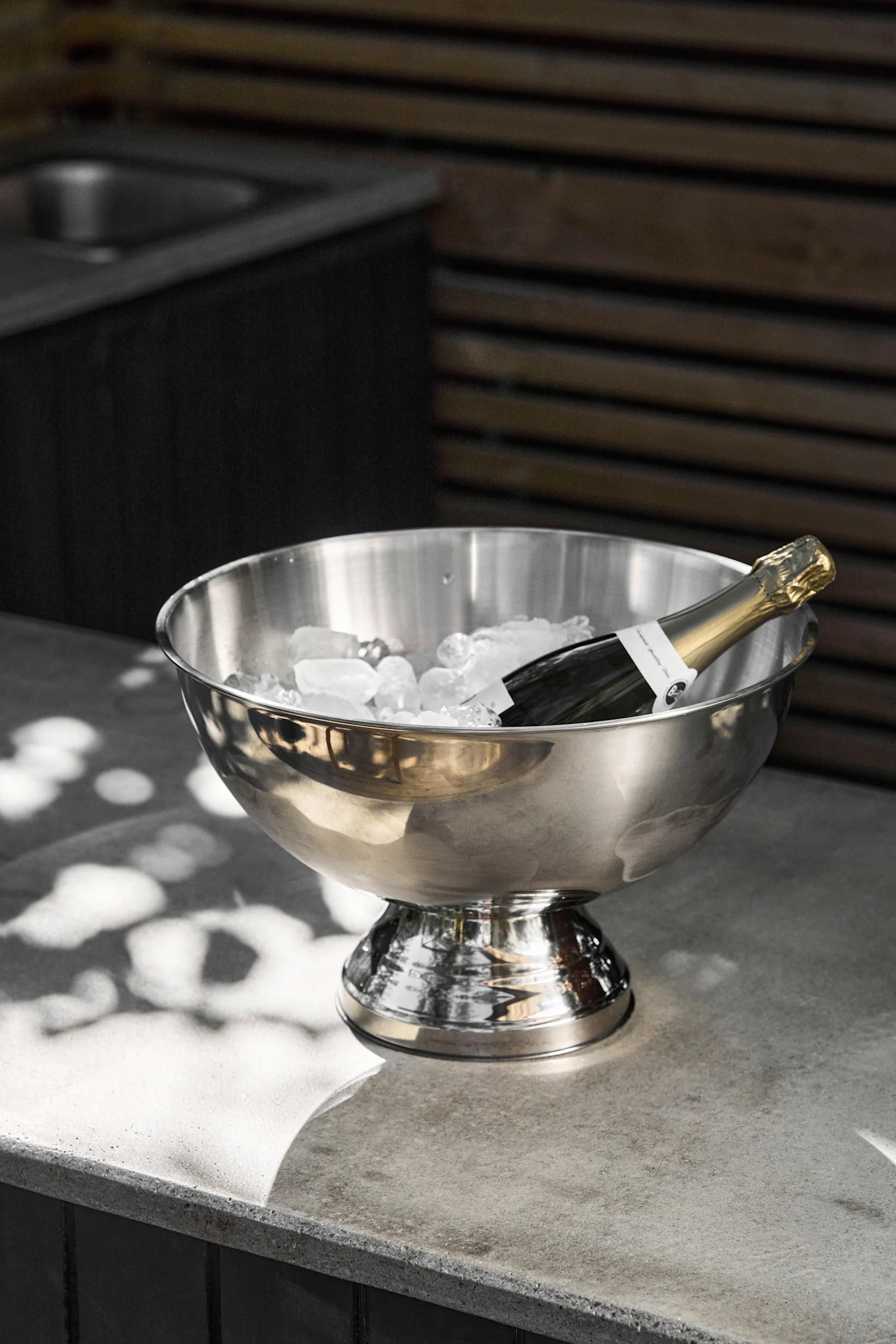 Una botella de champán reposa en una cubitera de acero inoxidable llena de cubitos de hielo sobre una encimera de hormigón gris, con luz solar moteada.