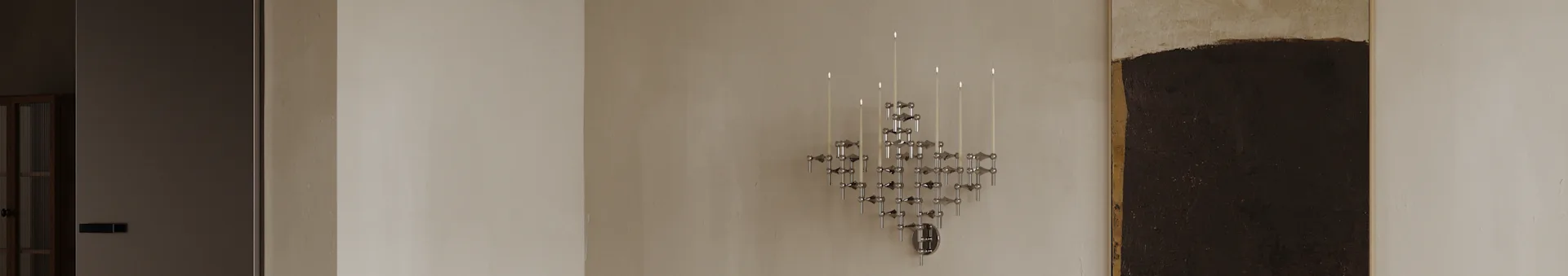 Un candelabro modular de cromo geométrico moderno con varias velas blancas encendidas montado en una pared clara.