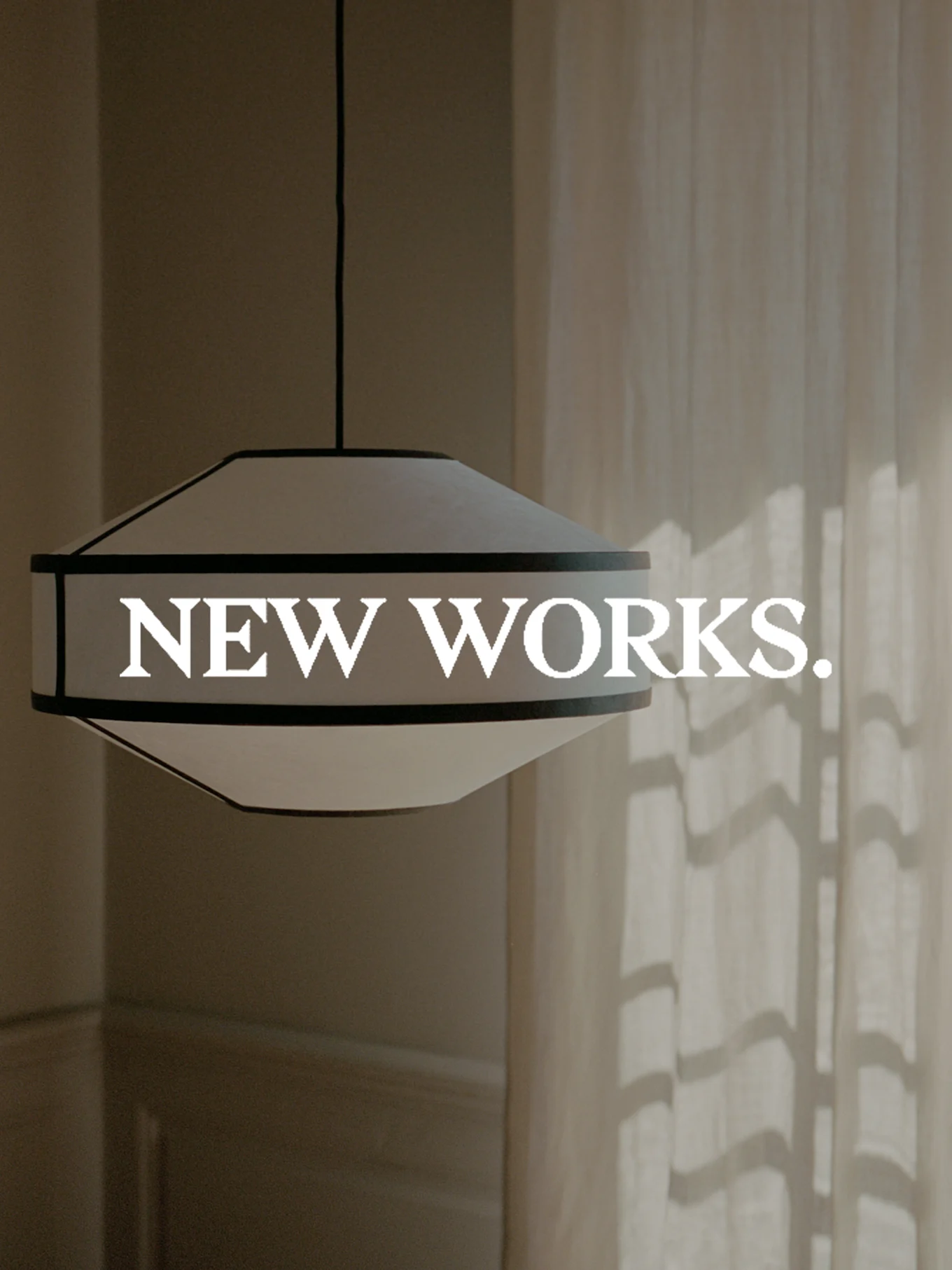 Una lámpara colgante ancha, blanca y con borde oscuro, cuelga con el texto 'NEW WORKS.' superpuesto. La luz del sol proyecta sombras sobre una cortina.
