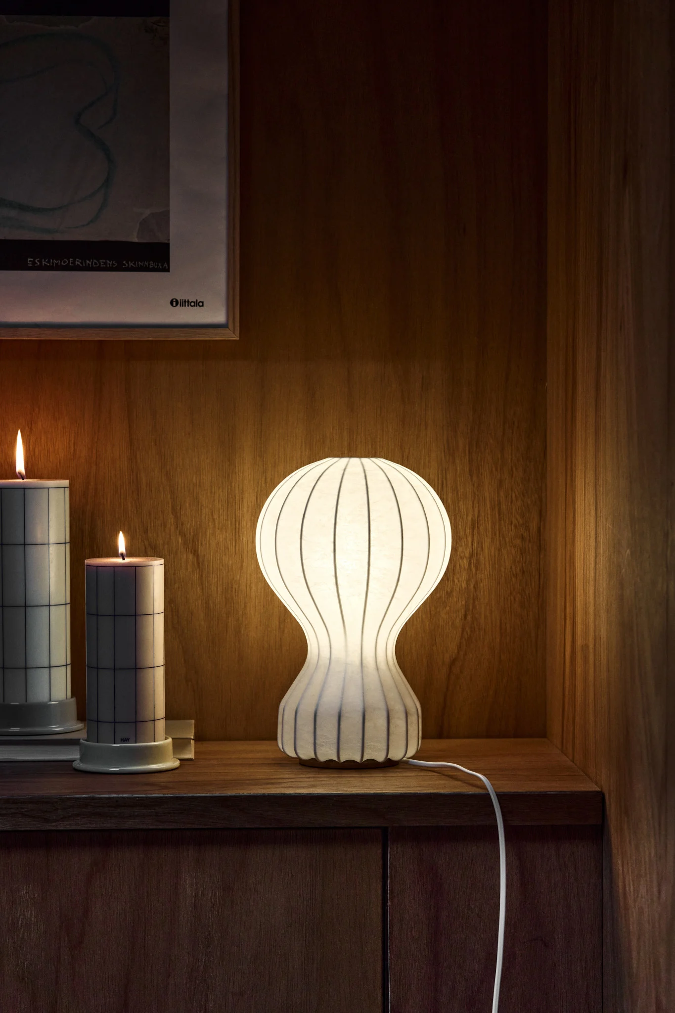 Lámpara de mesa blanca acanalada iluminada, dos velas con patrón de rejilla y arte en un estante de madera.