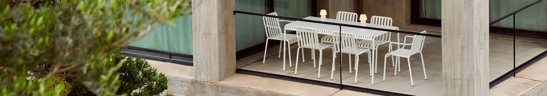 Un conjunto de comedor exterior con una mesa blanca de lamas y seis sillas a juego, iluminado por tres lámparas portátiles, en un patio de hormigón con plantas.