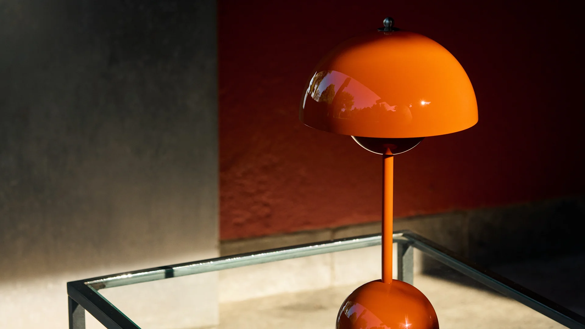 Lámpara de mesa brillante naranja con forma de seta sobre una mesa de cristal contra una pared roja y gris, con luz solar moteada.