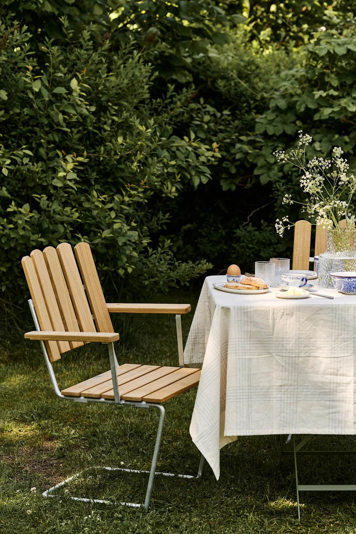 Mesa de jardín con mantel blanco, platos de desayuno y silla de listones de madera sobre césped verde.