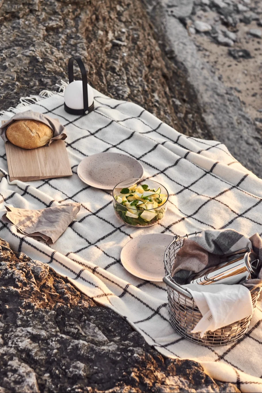 Picnic en rocas con manta a cuadros, pan, linterna, ensalada, platos y cesta.