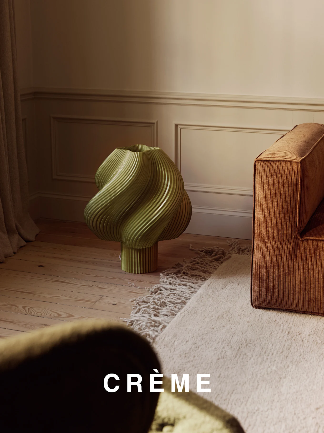 Una lámpara verde claro, retorcida y acanalada, sobre un suelo de madera, junto a un sofá acanalado marrón y una alfombra peluda beige.