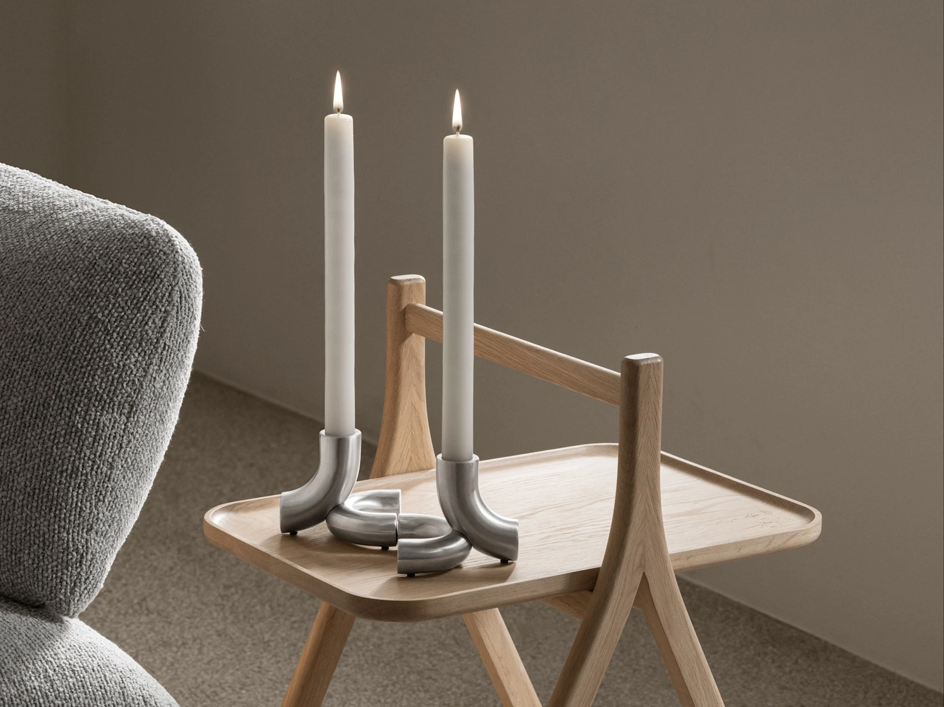 Dos velas blancas encendidas en candelabros plateados curvos sobre una mesa auxiliar de madera clara, con un sillón gris a la izquierda.