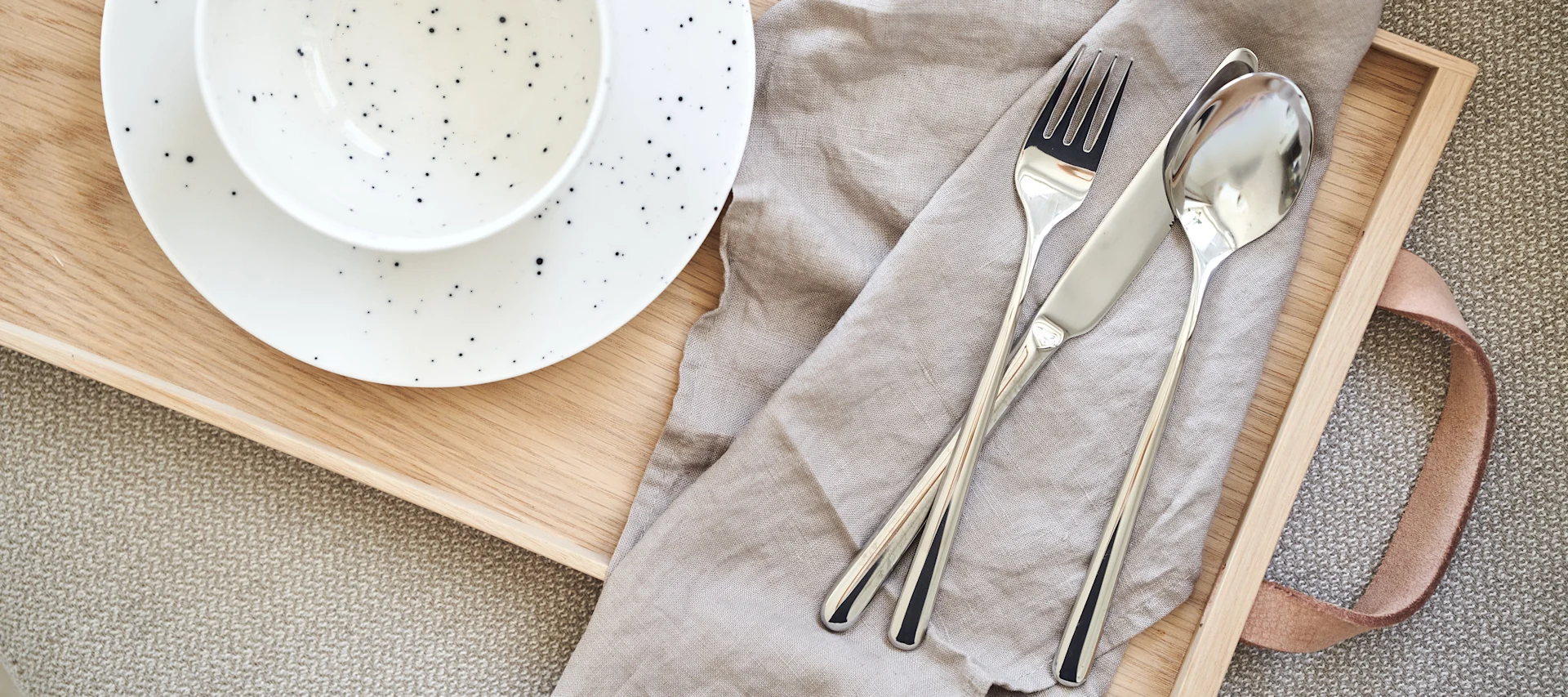 Vajilla con plato y cuenco blancos salpicados sobre una bandeja de madera, junto a un tenedor, cuchillo y cuchara sobre una servilleta de lino beige.