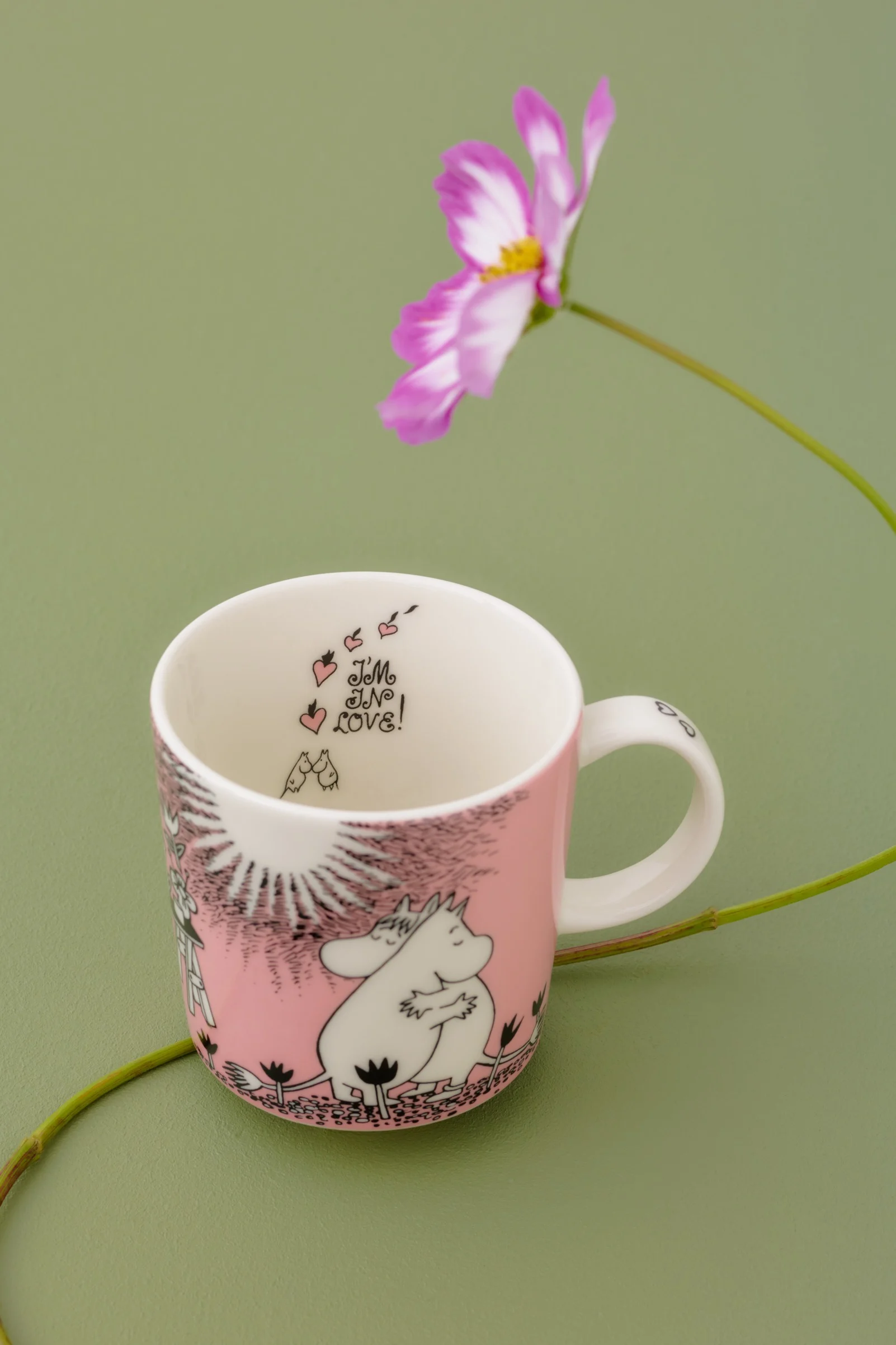 Taza Moomin rosa y blanca con Moomintroll y Snorkmaiden, "I'M IN LOVE!" dentro, sobre fondo verde con flor.