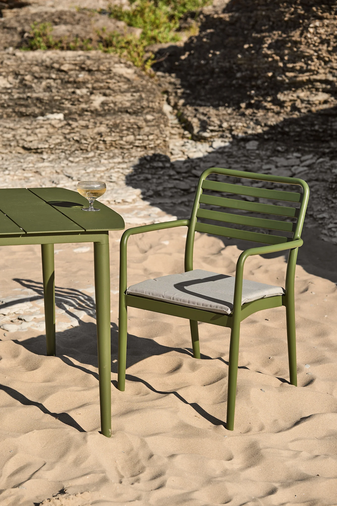 Mesa y silla de metal verde para exterior en una playa de arena con una copa de vino blanco en la mesa.
