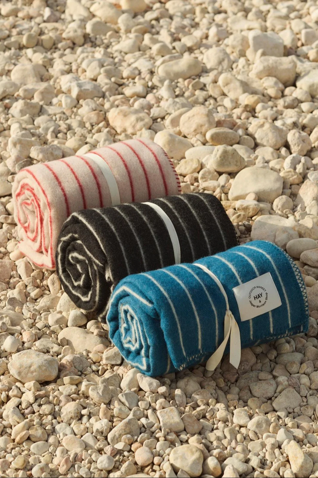 Tres mantas rayadas enrolladas en rosa, negro y azul, atadas con lazos en una playa de guijarros.