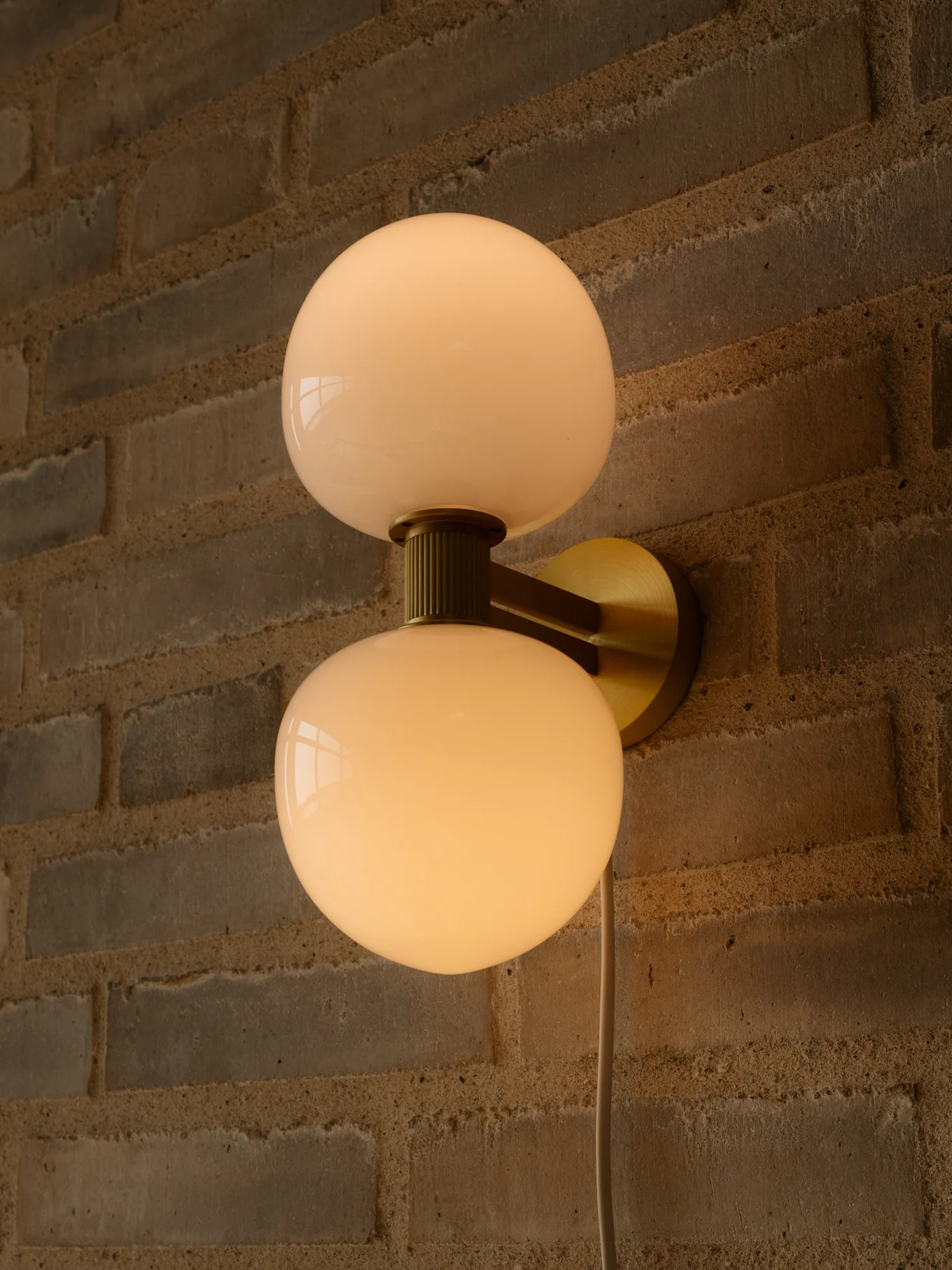 Lámpara de pared con dos pantallas de cristal esféricas blancas lechosas y soporte de latón en una pared de ladrillo.