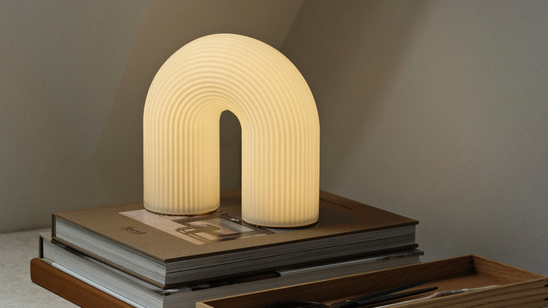 Lámpara de mesa arqueada con textura acanalada, brillando cálidamente sobre una pila de libros marrones, con una bandeja de madera cerca.