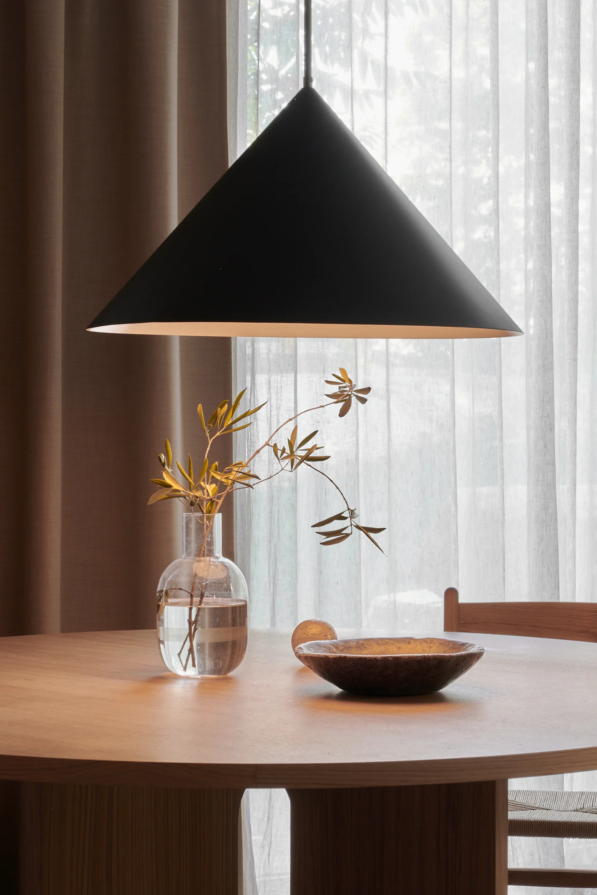 Mesa de comedor redonda de madera con una lámpara colgante cónica negra encima, con un jarrón transparente con ramas y un cuenco de madera oscuro.
