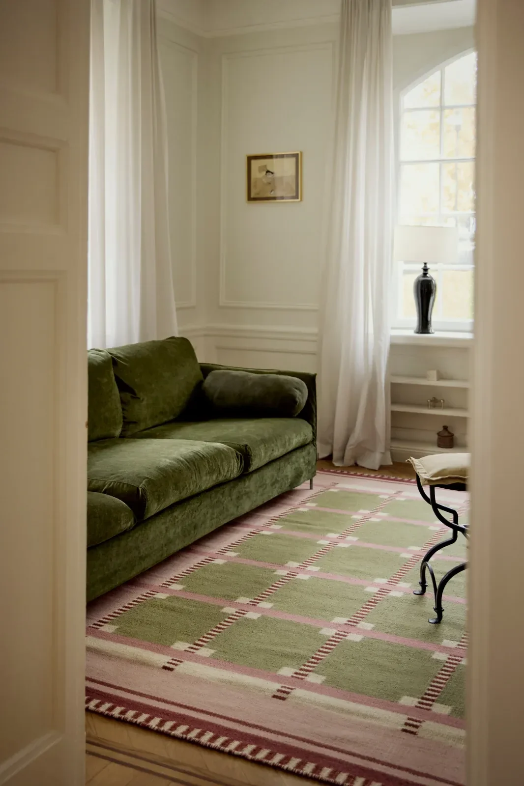 Una sala de estar con un sofá de terciopelo verde, una alfombra estampada verde y rosa, y una ventana con cortinas blancas.
