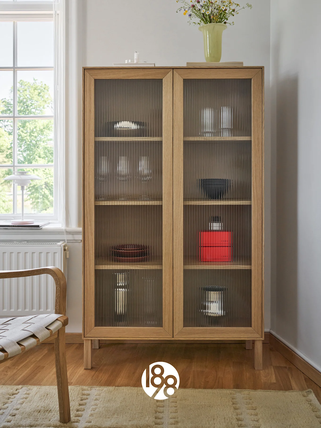 Un armario de madera clara con puertas de cristal estriado que exhibe platos y vasos, coronado con un jarrón de flores silvestres. Una silla y una ventana están a la izquierda.