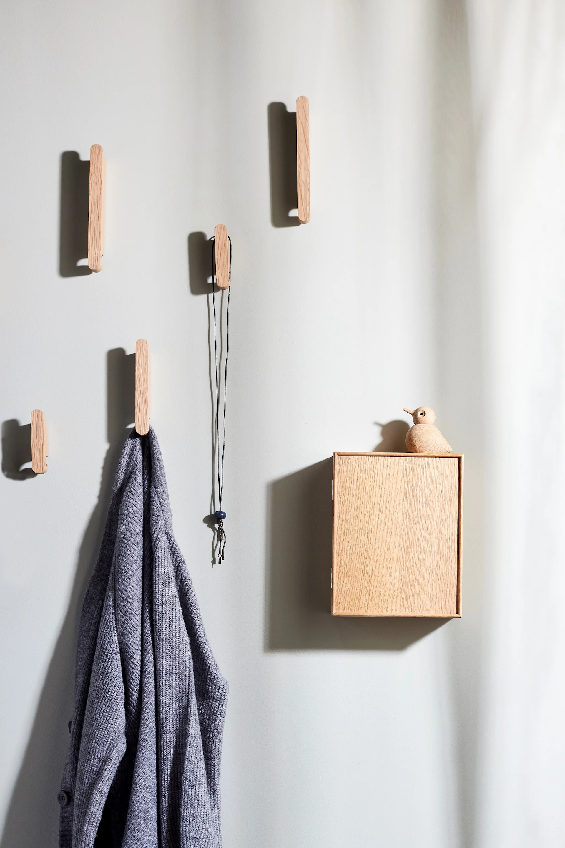 Percheros de madera, un cárdigan gris y una caja de madera con una figura de pájaro en una pared clara.