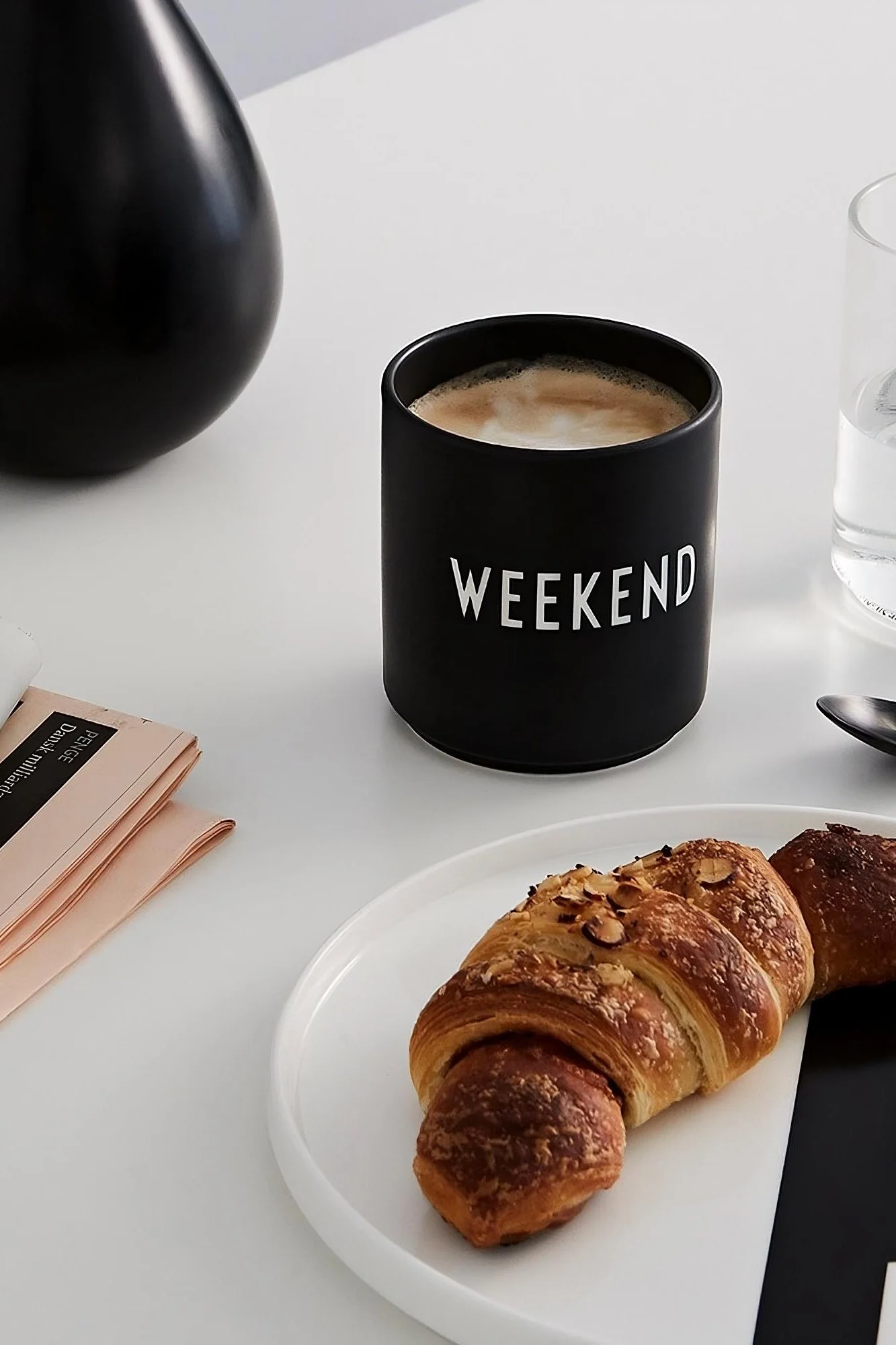 Taza de café: la taza de café negra con la impresión "Weekend" de Design Letters está sobre una mesa, frente a ella un plato con un croissant.