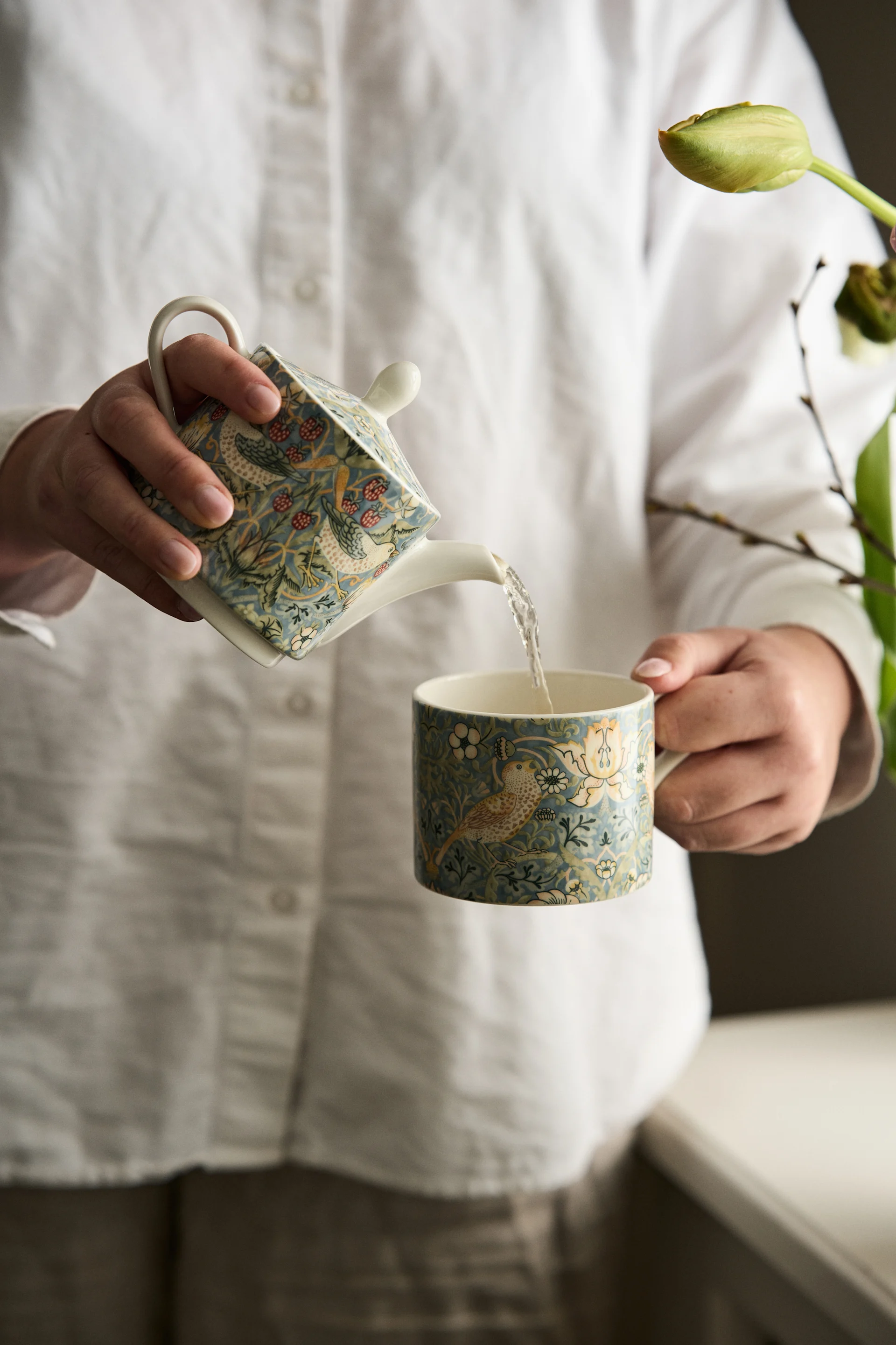 Persona con camisa blanca vertiendo agua de una tetera decorativa con patrón de pájaros y bayas en una taza a juego.