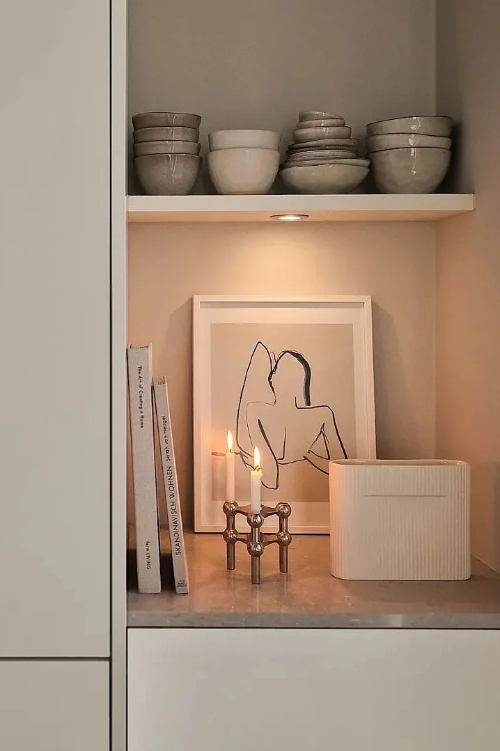 Un estante acogedor con cuencos de cerámica, libros, arte enmarcado, velas encendidas en un portavelas de latón y un jarrón estriado.