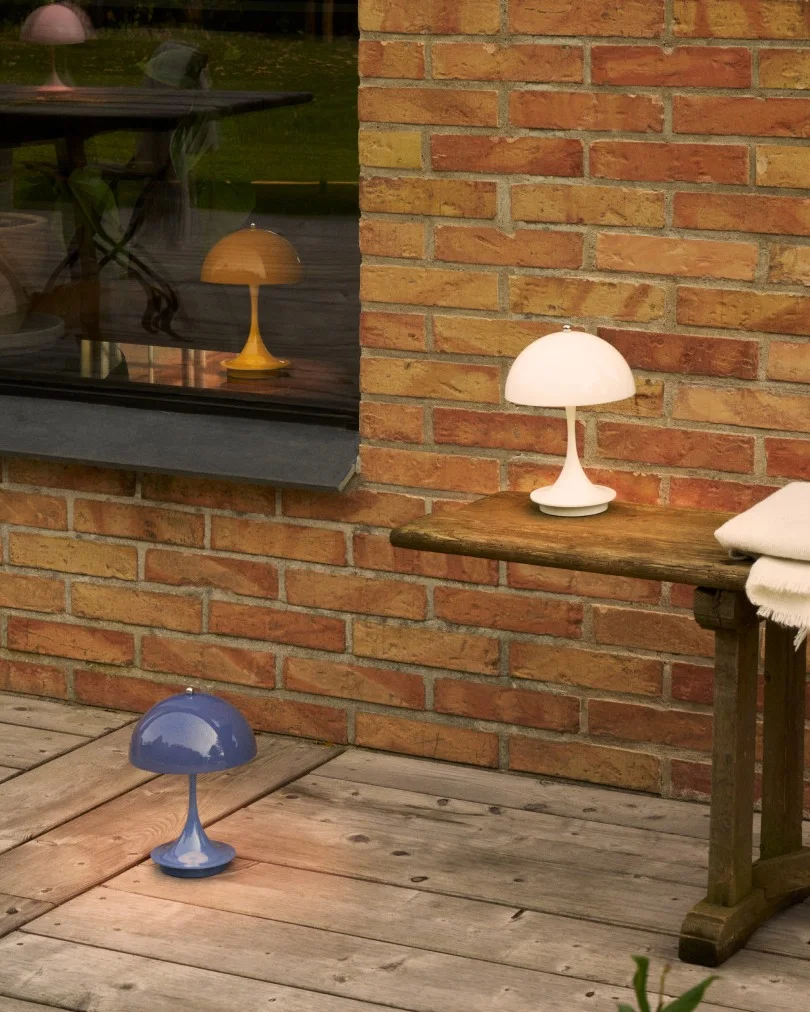 Tres lámparas con forma de seta en blanco, azul y amarillo en un patio de madera junto a una pared de ladrillo y ventana.