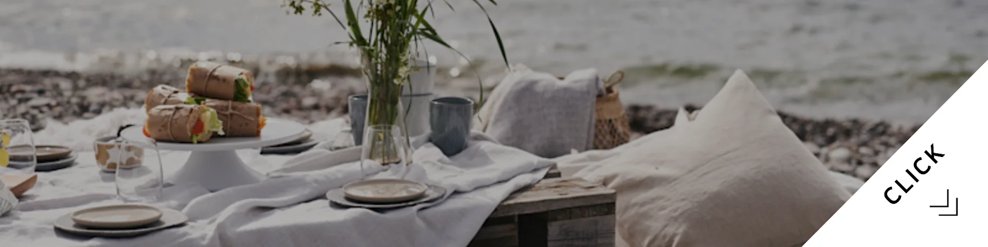 Un tranquilo picnic en la playa con comida envuelta, vajilla, un jarrón de flores y cojines en una orilla de guijarros junto al mar.