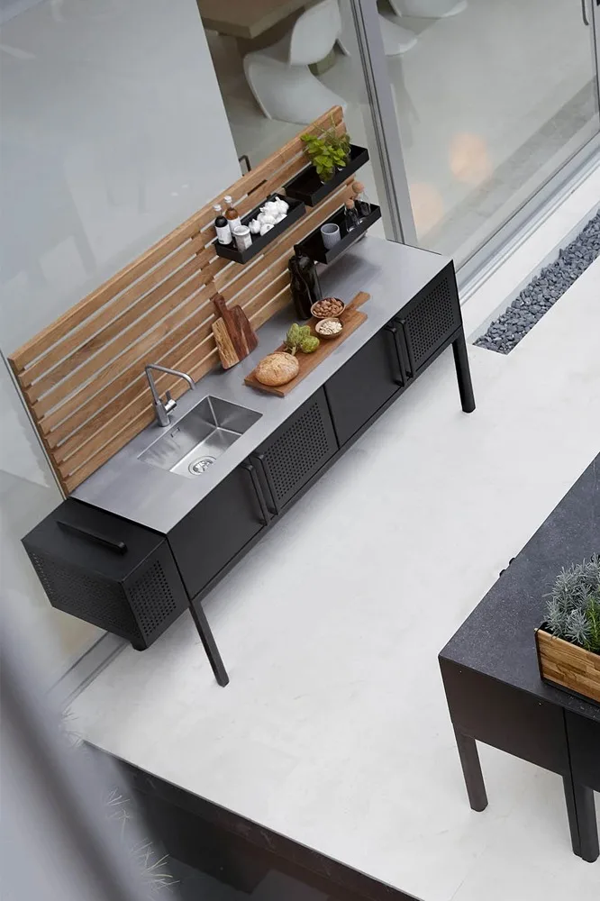 Moderna cocina exterior con fregadero de acero inoxidable, armarios negros, panel trasero de madera y estantes con especias y comida.