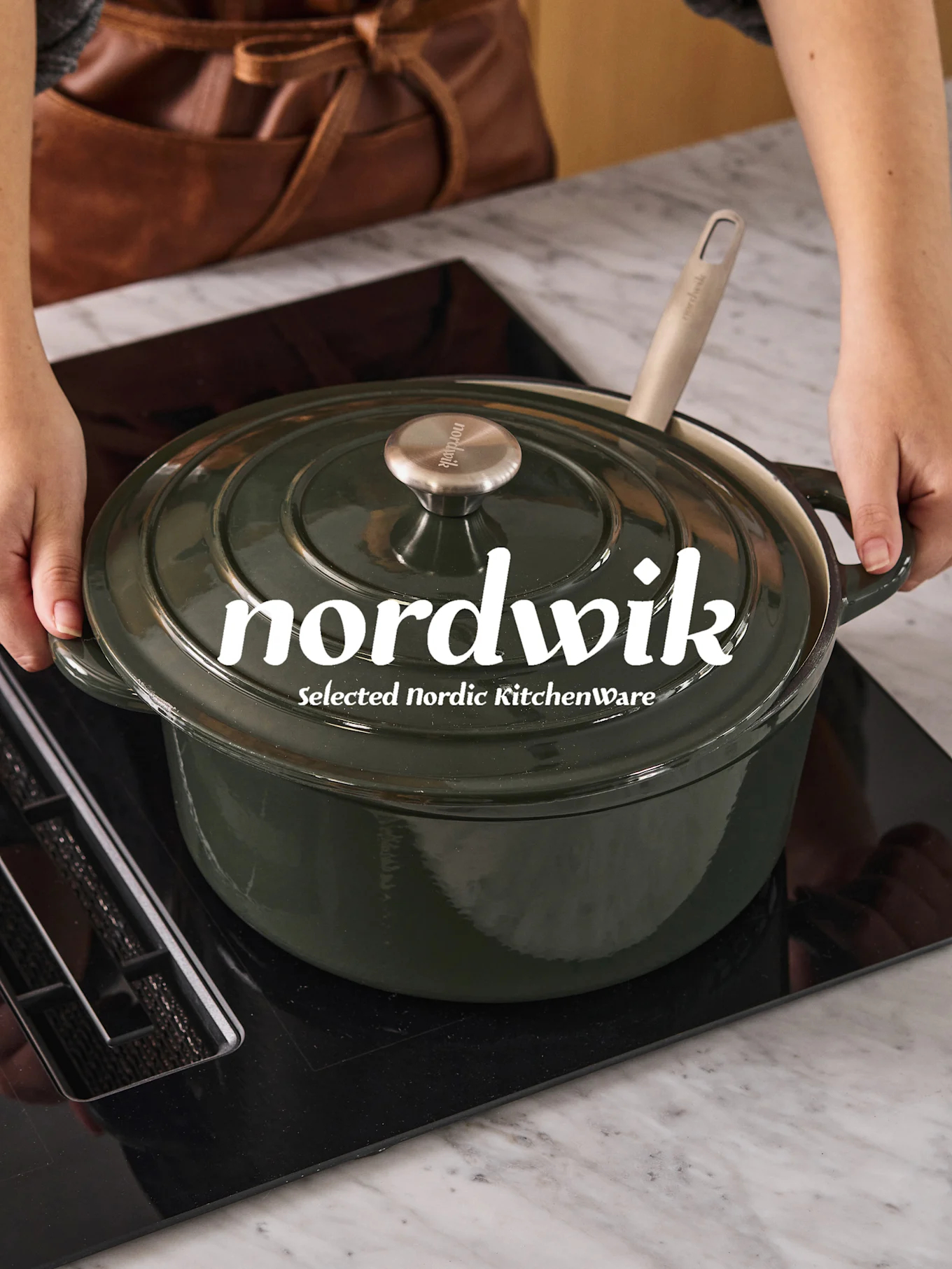 Manos colocando una olla de hierro fundido verde oscuro con una cuchara clara en una placa de inducción negra. 'nordwik' es visible en la tapa.