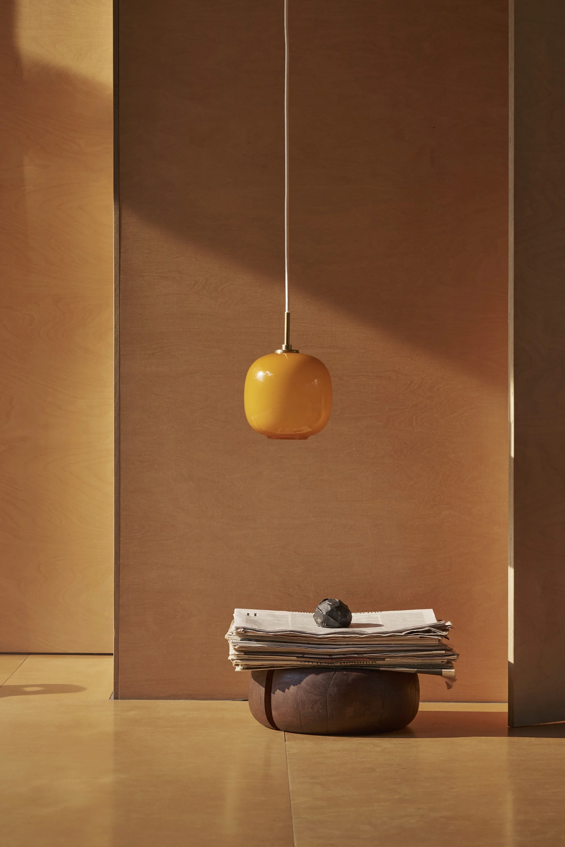 Una lámpara colgante ovalada naranja cuelga sobre una pila de periódicos y un objeto decorativo oscuro sobre una base de madera en una habitación minimalista.