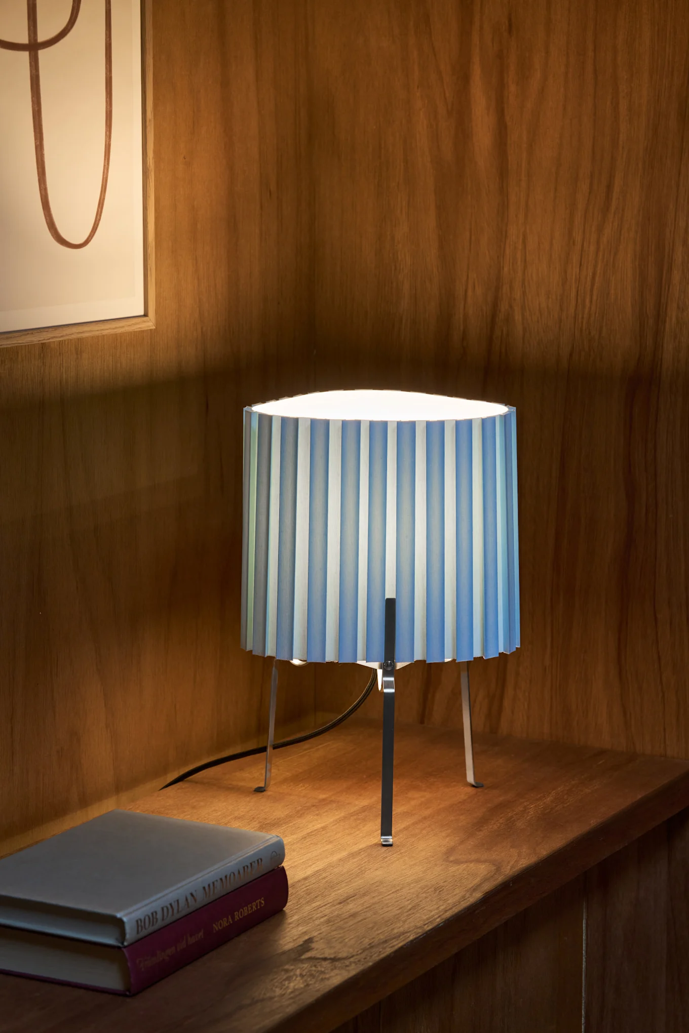 Una lámpara de mesa con una pantalla plisada de rayas azules y blancas y patas de metal, sobre un estante de madera junto a dos libros.