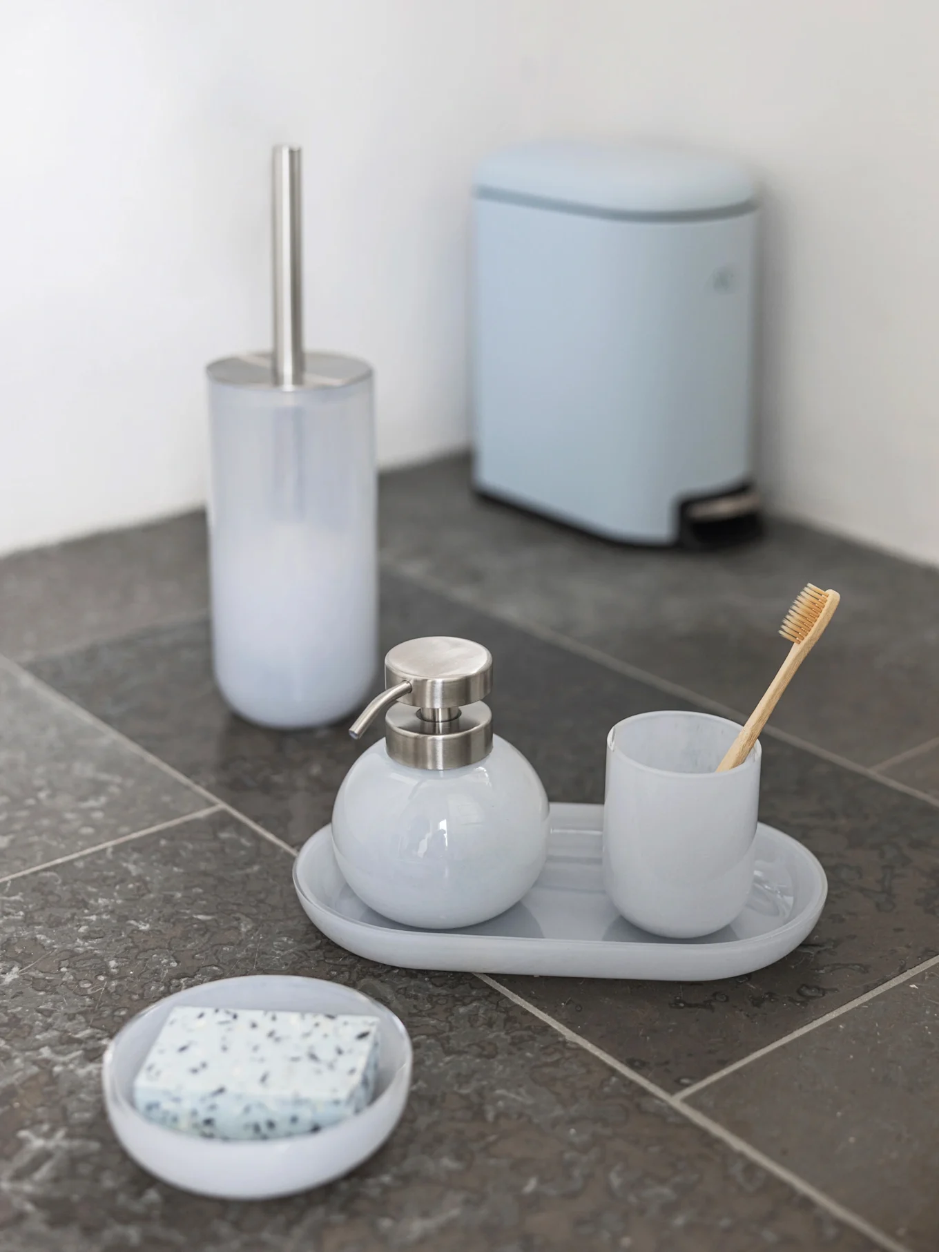 Set de baño azul claro: escobilla, dispensador de jabón, vaso con cepillo de dientes de bambú, jabonera y cubo de pedal sobre azulejos oscuros.