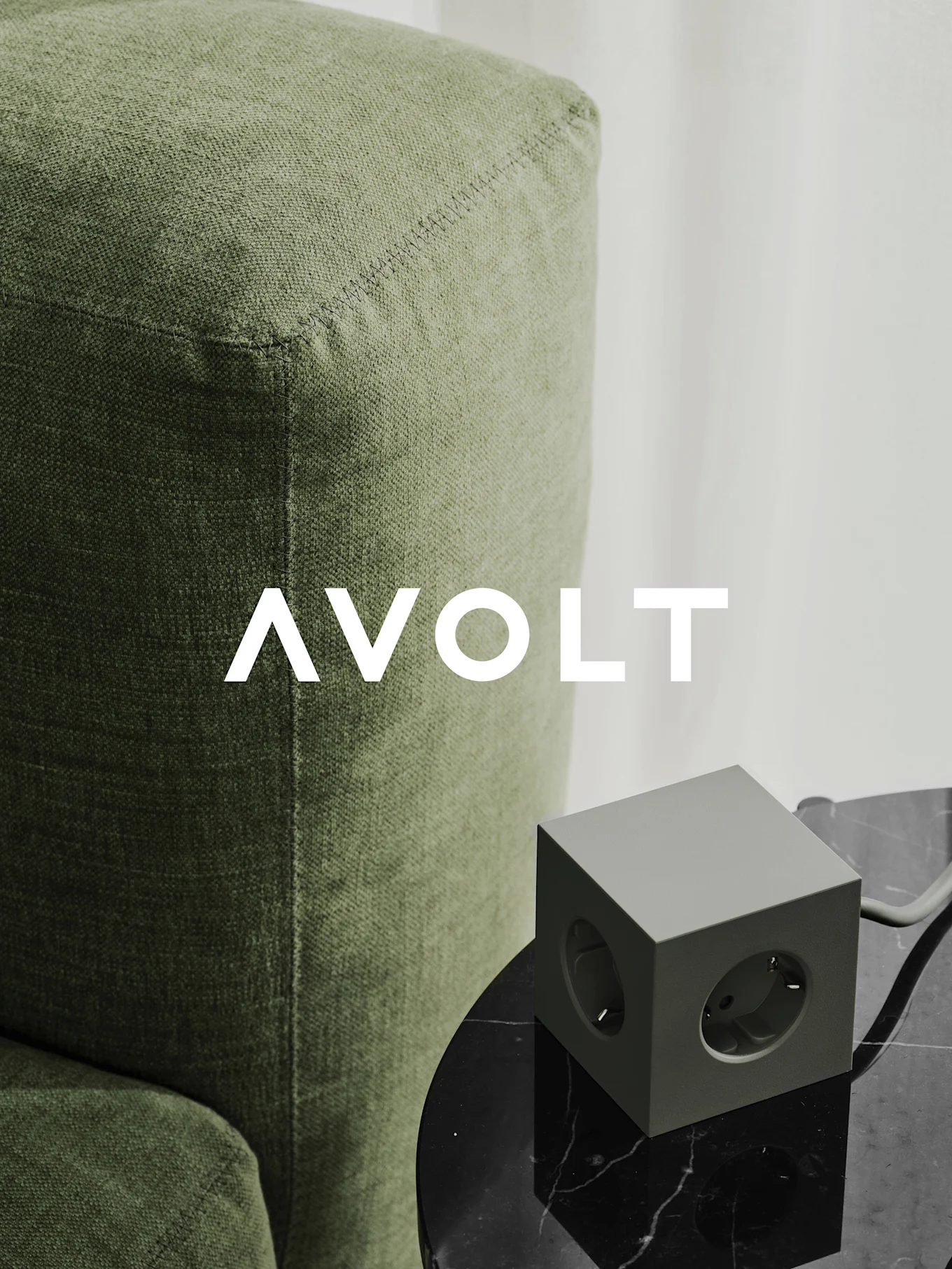 Una regleta verde en forma de cubo con tres tomas en una mesa de mármol negro junto a un sofá verde. Logo de AVOLT.