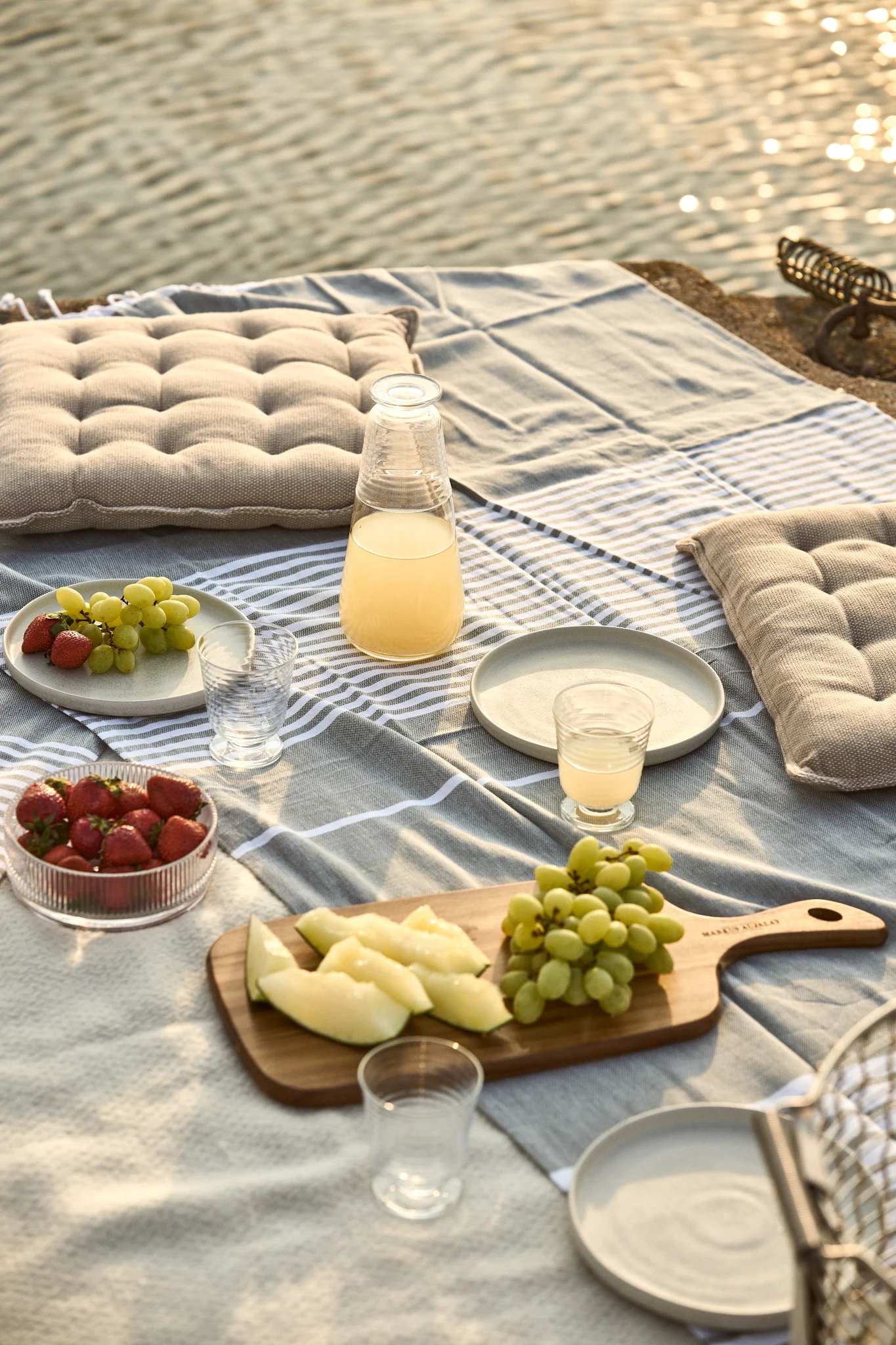 Picnic junto al agua con manta de rayas, cojines, zumo, fresas, uvas y rodajas de melón.