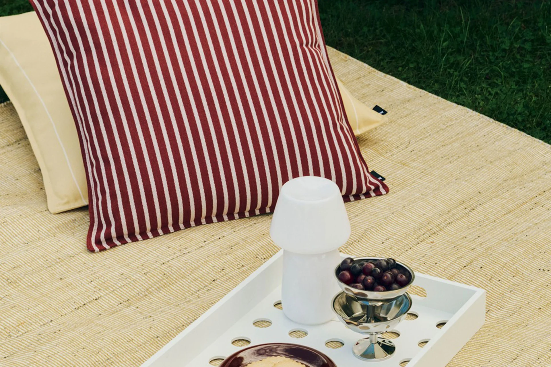 Montaje de picnic al aire libre con cojín a rayas rojas y blancas, cojín amarillo, bandeja blanca con lámpara de seta y un cuenco de bayas.
