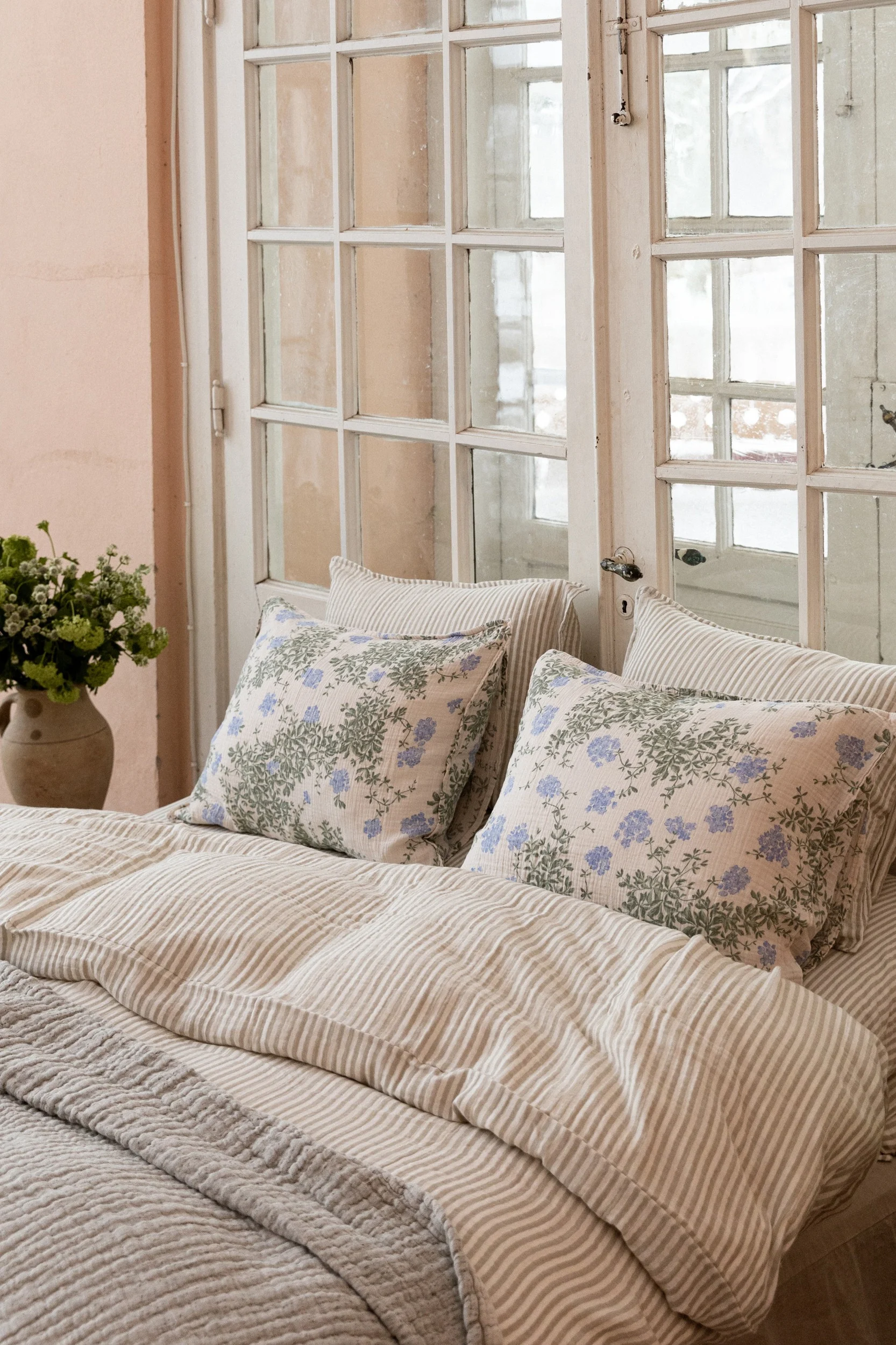 Una cama con edredón a rayas y dos almohadas con estampado floral azul, frente a una ventana con paneles. Un jarrón con follaje verde a la izquierda.