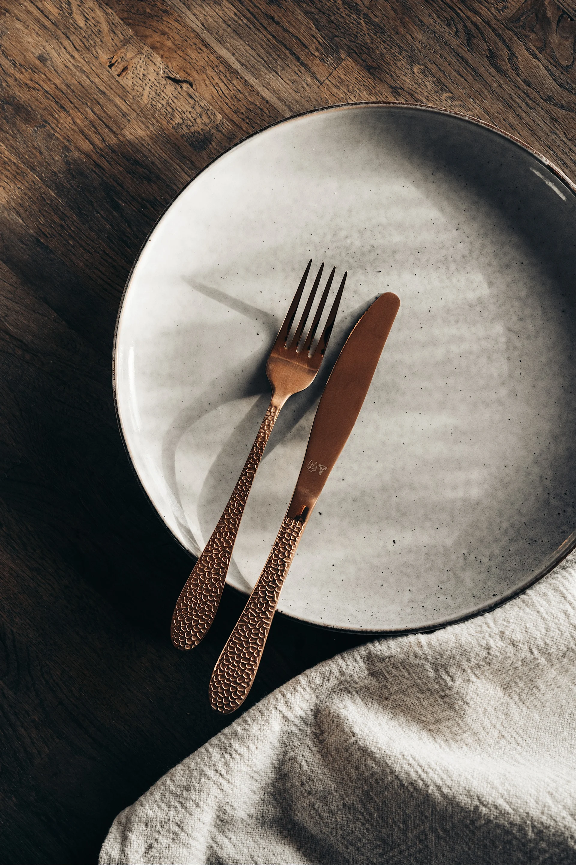 Tenedor y cuchillo color cobre con mangos texturizados sobre un plato de cerámica gris claro, junto a una servilleta de lino sobre madera.